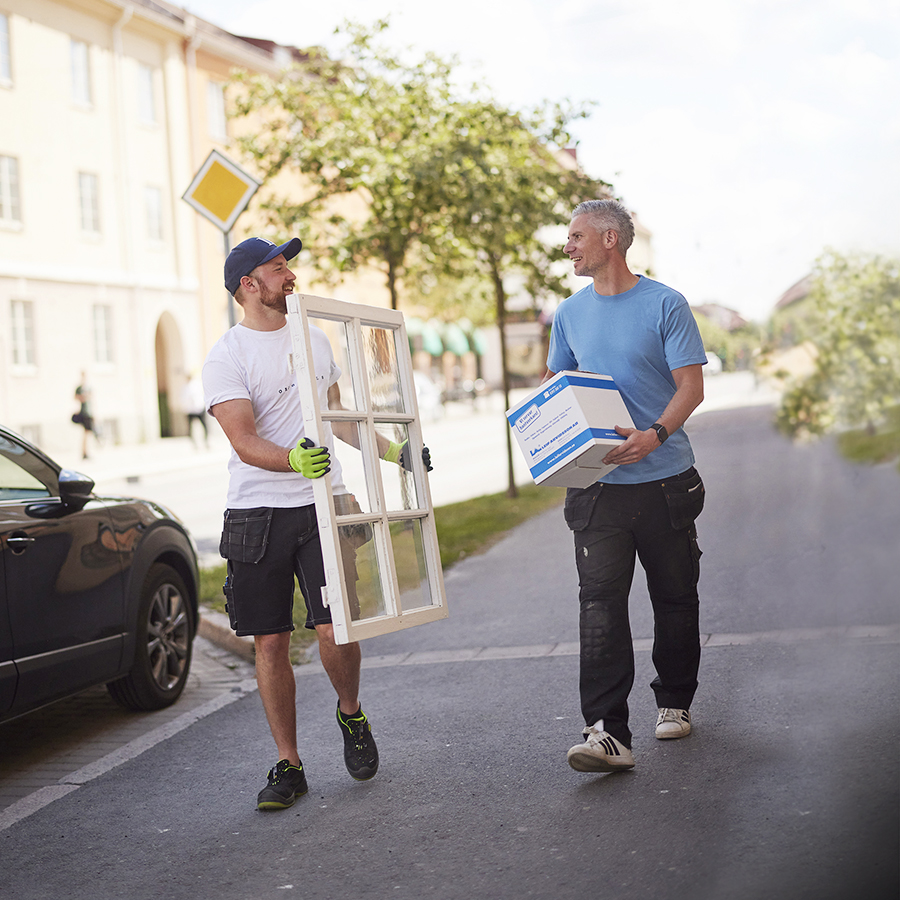 Två hantverkare går på en trottoar, den ene håller i ett fönster och den andre i ett paket från Leif Arvidsson AB